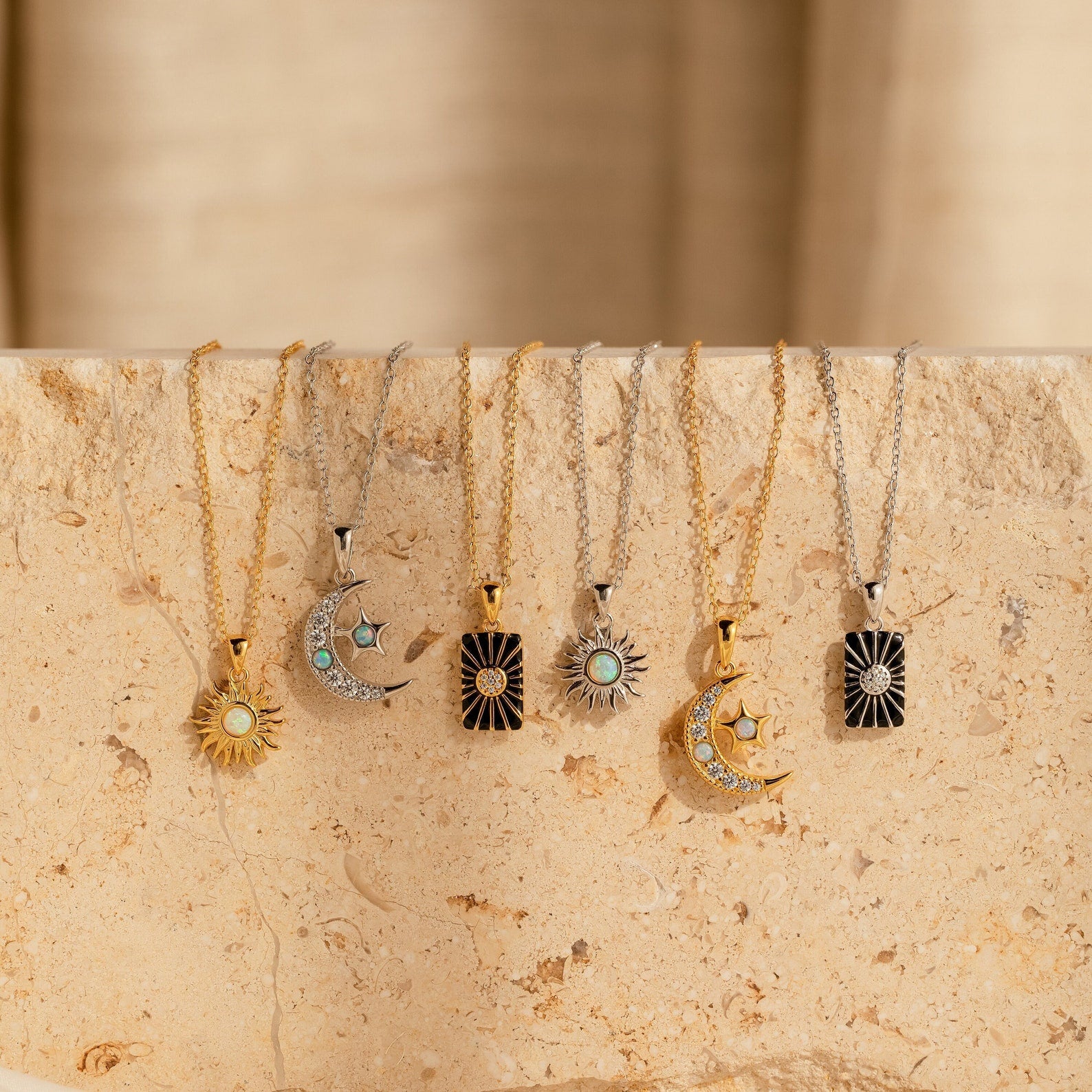 Six gold and silver Celestial Pendant Necklace Set pieces with sun, moon, and star charms—some featuring opal and diamond accents—are arranged on a beige stone surface. A perfect gift for her, inspired by the beauty of the night sky.
