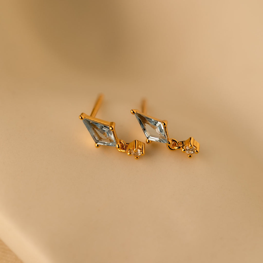 Aquamarine Kite Drop Earrings with diamond-shaped blue CZ gemstones and small square accents on gold studs, displayed on a beige background.