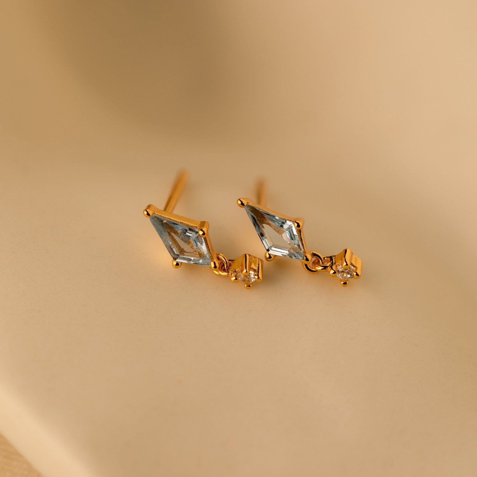 Aquamarine Kite Drop Earrings with diamond-shaped blue CZ gemstones and small square accents on gold studs, displayed on a beige background.