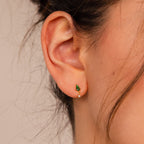 Close-up of a person's ear wearing a small gold earring with a green stone and tiny dangling bead, similar to the elegant Emerald Kite Drop Earrings.
