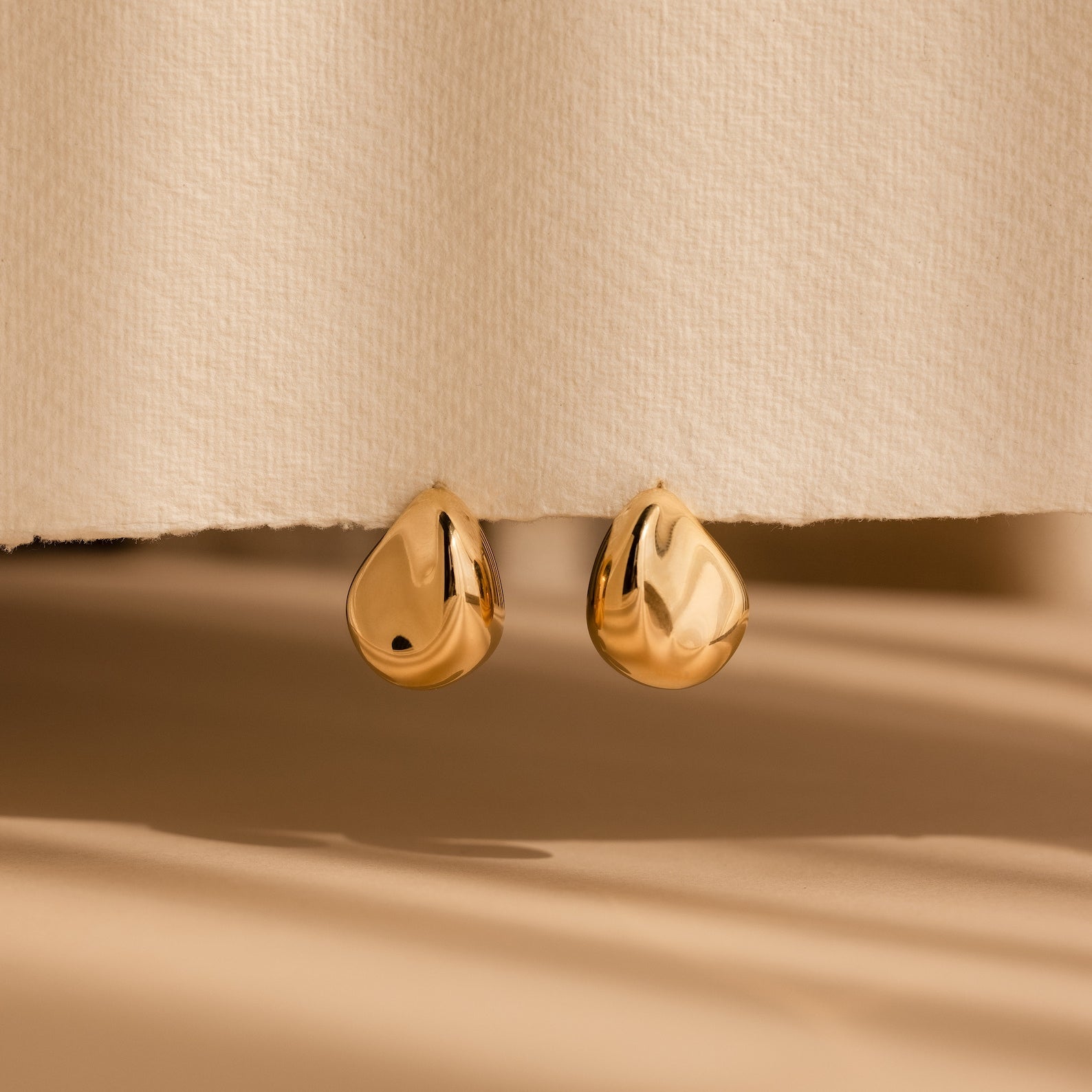 A pair of Bold Teardrop Studs in shiny gold, shown against a textured beige background—perfect statement earrings for any occasion.