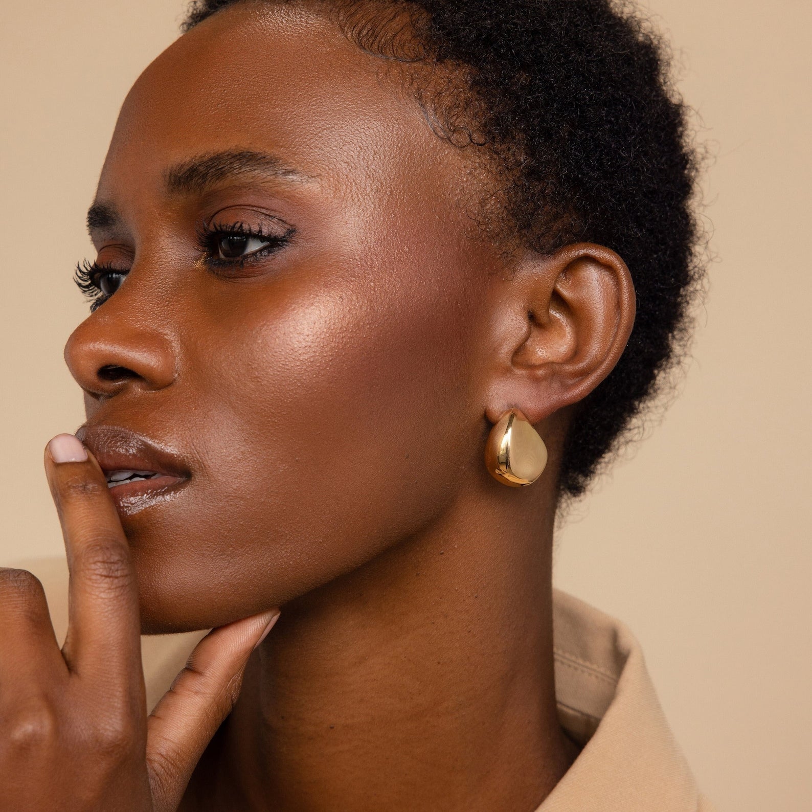 A woman with short curly hair wears Bold Teardrop Studs and touches her lips, gazing thoughtfully to the side.