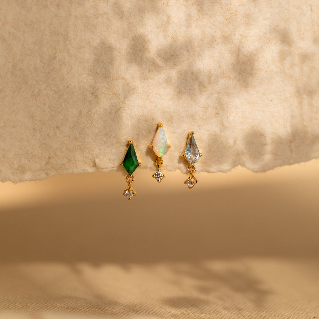 Three gold Emerald Kite Drop Earrings featuring green, opal, and blue stones are elegantly displayed on a textured beige surface—a stunning addition to any jewelry collection.