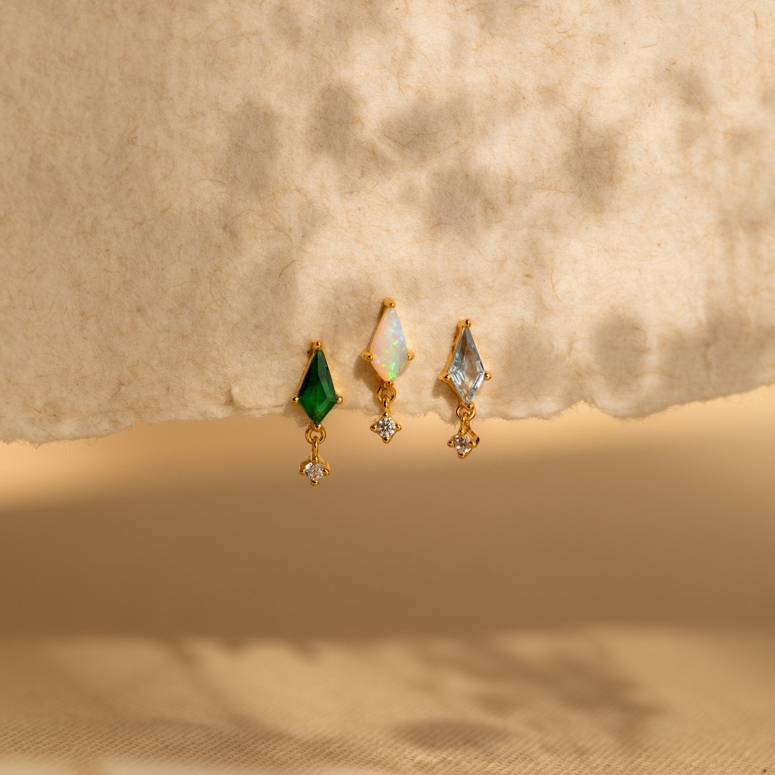 Three gold Emerald Kite Drop Earrings featuring green, opal, and blue stones are elegantly displayed on a textured beige surface—a stunning addition to any jewelry collection.