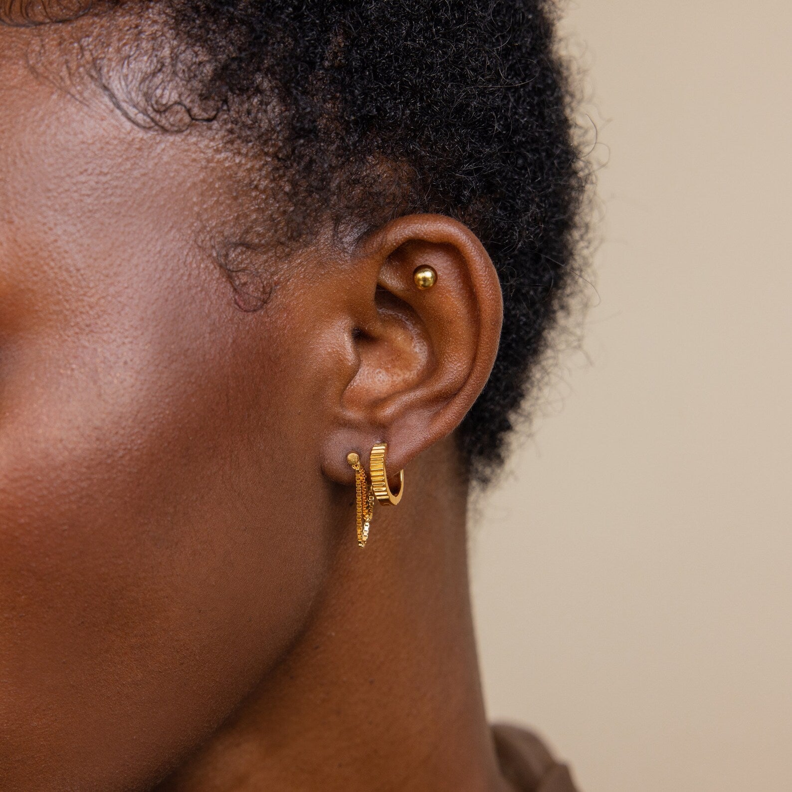 Close-up of a person’s ear with a stylish earring stack: a stud, two small hoops, and the elegant Flatback Chain Earrings in gold.