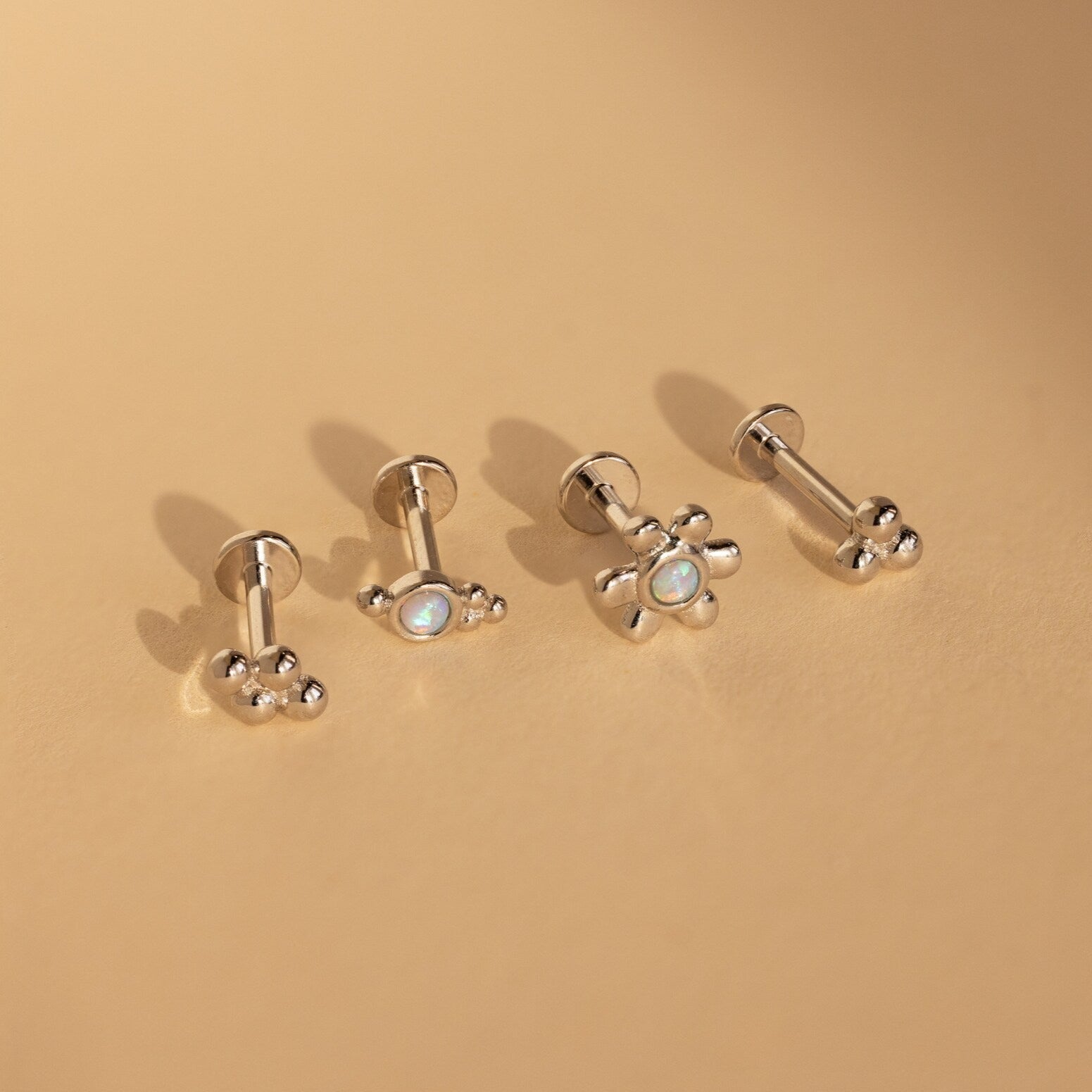 The Opal Cluster Studs Set includes four silver stud earrings with flower and paw designs, displayed on a beige surface—ideal accents for cartilage piercings.