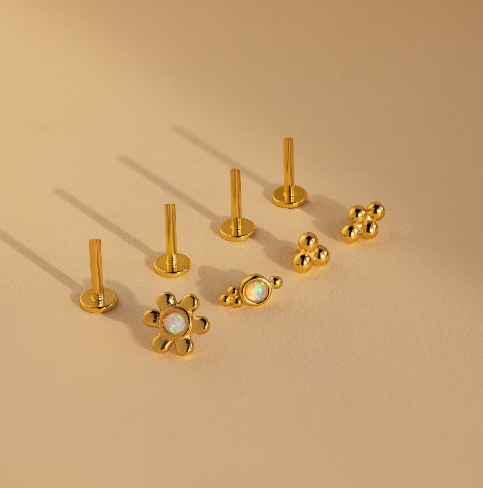 The Opal Cluster Studs Set includes five gold earrings with unique shapes, perfect for cartilage piercings, featuring floral and beaded designs, all displayed on a beige background.