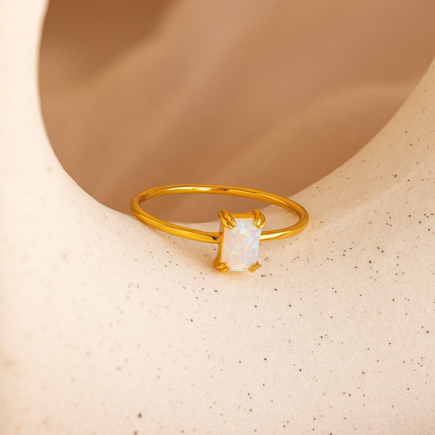 The Opal Emerald Ring features a rectangular opal set in gold, shown on a textured beige background—an elegant anniversary gift choice.