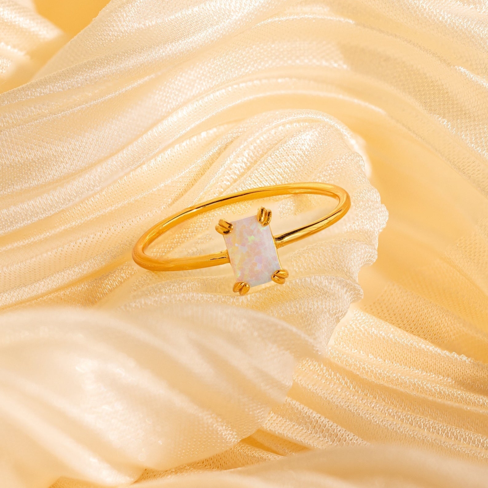 The Opal Emerald Ring, featuring a rectangular opal stone, rests on soft cream satin fabric—an elegant anniversary gift.
