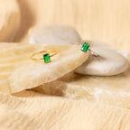 The Harley Emerald Ring, with its vibrant green rectangular gemstone in gold or silver, evokes vintage art deco style. Displayed on smooth beige stones, each ring radiates timeless elegance and sophisticated charm.