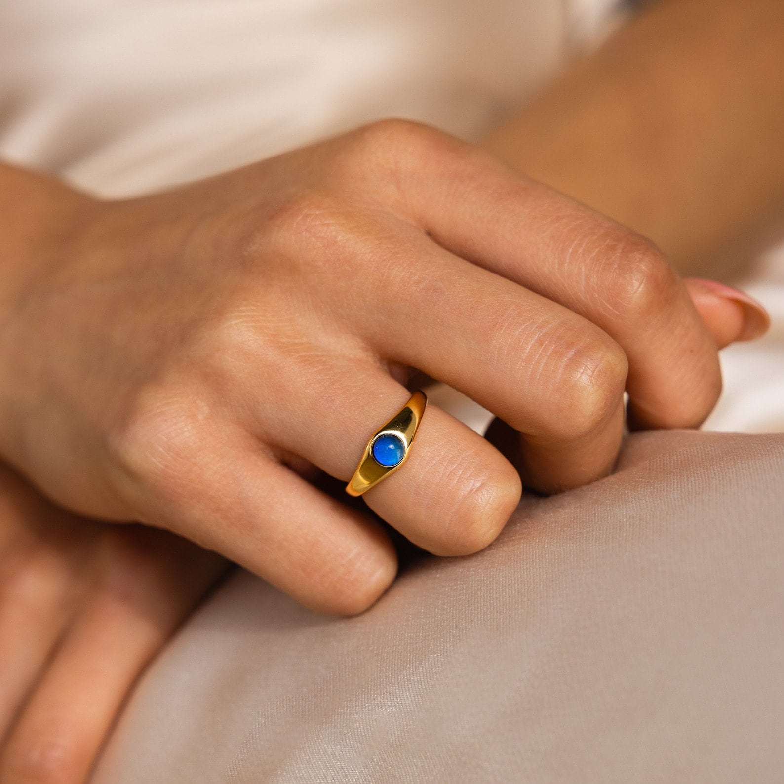 A hand wearing a gold ring from the Signet Mood Rings Set, featuring a round blue stone, rests on light fabric and captures the nostalgic charm of classic jewelry.
