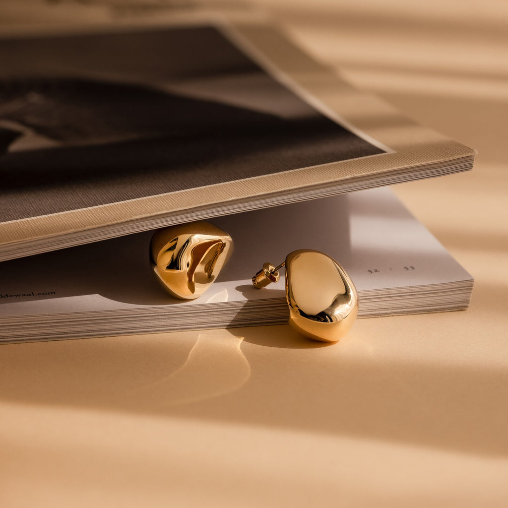 The Bold Teardrop Studs, two shiny gold statement earrings, rest on a slightly open magazine bathed in warm, soft light.