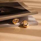 The Bold Teardrop Studs, two shiny gold statement earrings, rest on a slightly open magazine bathed in warm, soft light.