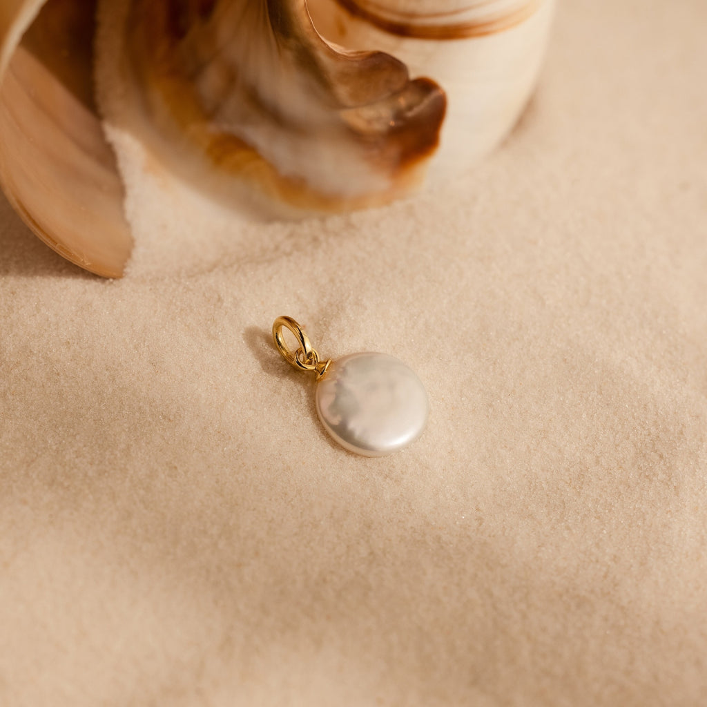 Close up of a single round coin shaped freshwater pearl charm with a gold plated bail, photographed in natural light on soft sand with seashells in the background.