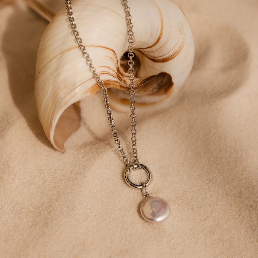 The Round Pearl Charm in Sterling Silver necklace lies on sand next to a spiral seashell.