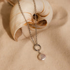 A Round Pearl Charm necklace rests on sandy ground beside a seashell, showcasing elegant jewelry inspired by the sea.