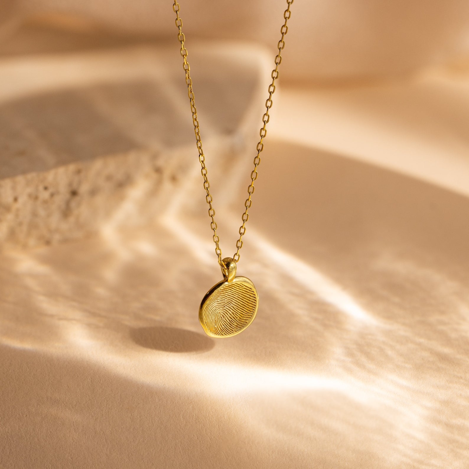 The Fingerprint Coin Necklace features a round gold pendant, shown on a beige surface with soft shadows—an ideal and meaningful Mother's Day gift.