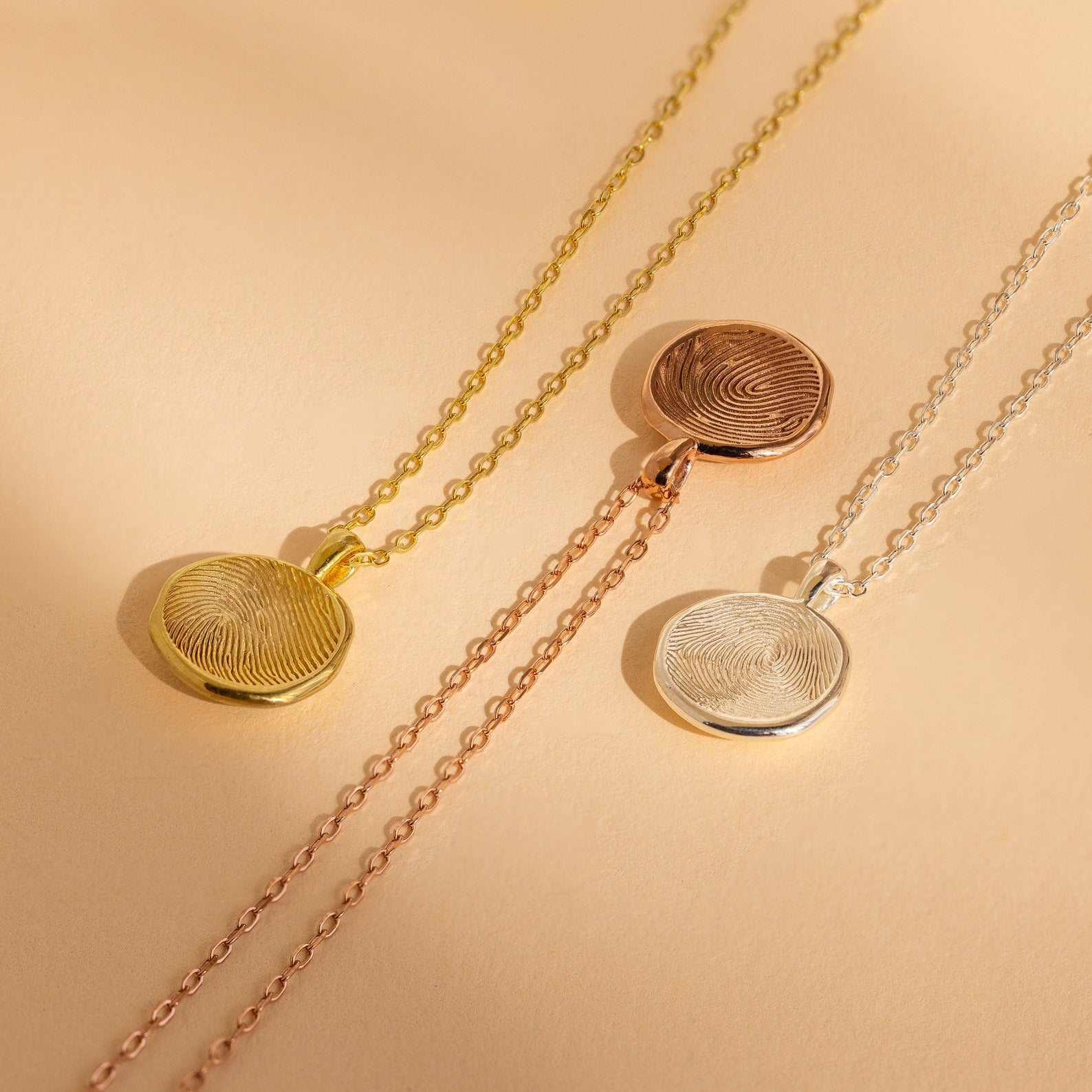 The Fingerprint Coin Necklace is shown on gold, rose gold, and silver chains against a beige background—a timeless piece perfect for gifting on Mother’s Day.