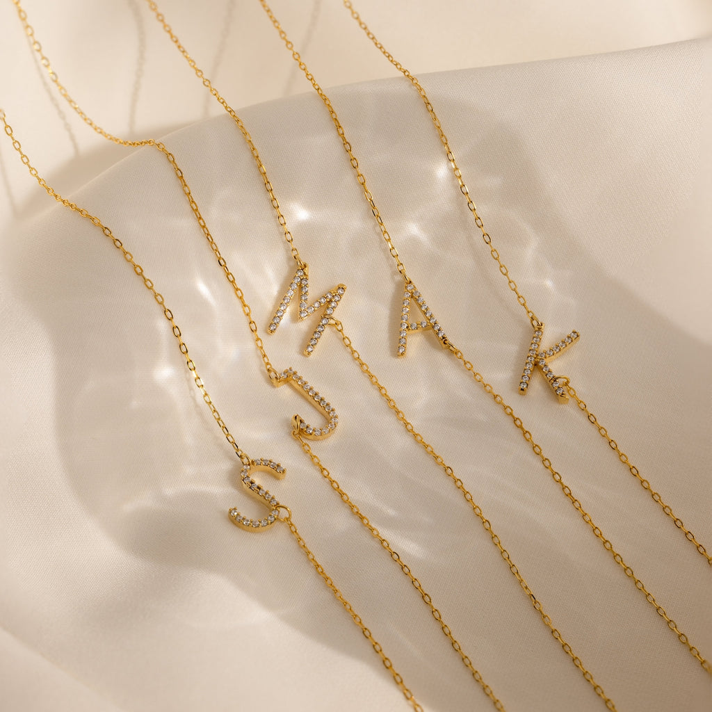 Four gold Mini Pave Sideways Initial Necklaces—featuring crystal-studded M, A, S, and K pendants—rest on soft beige fabric. Each personalized necklace adds a sparkling touch to your look.