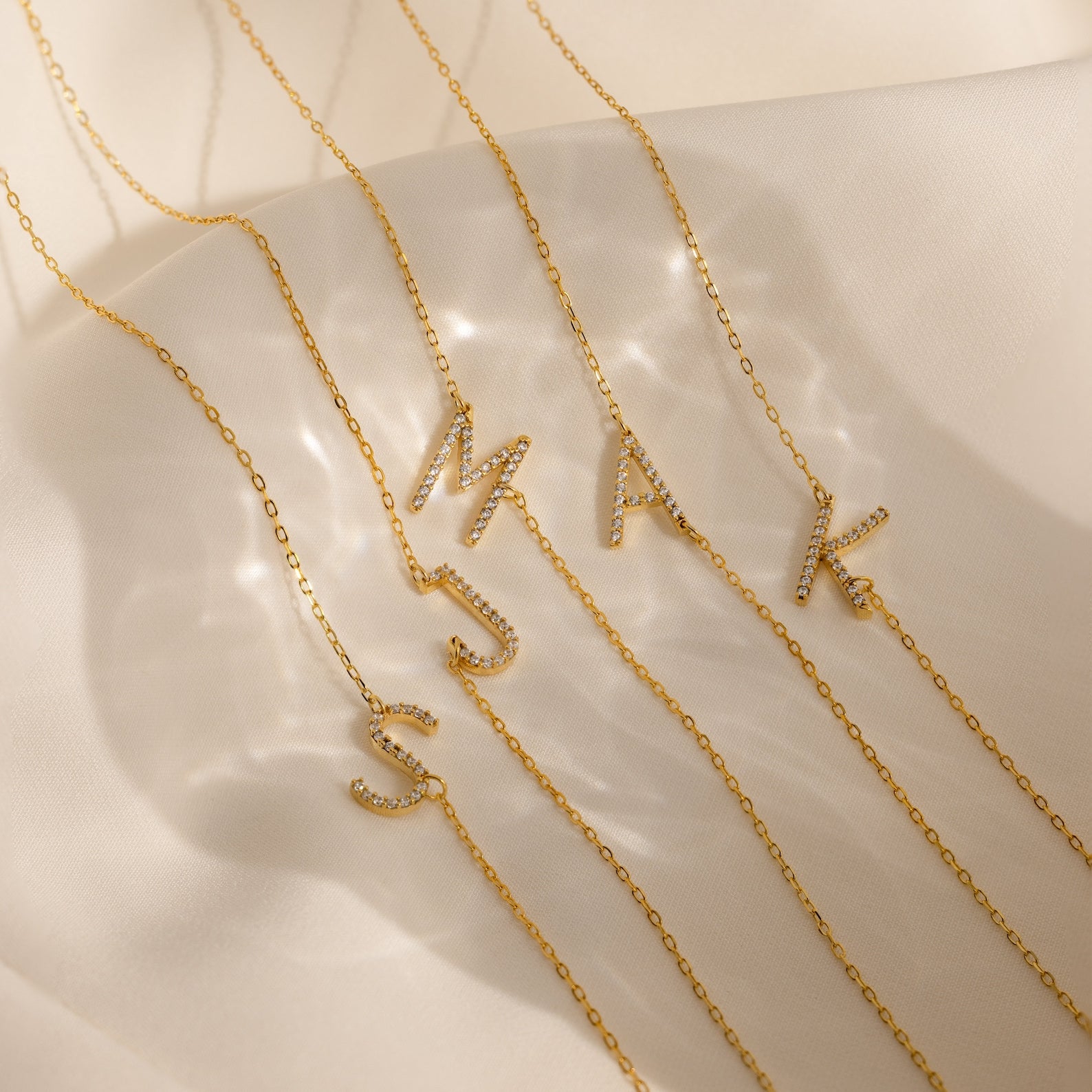 Four gold Mini Pave Sideways Initial Necklaces—featuring crystal-studded M, A, S, and K pendants—rest on soft beige fabric. Each personalized necklace adds a sparkling touch to your look.