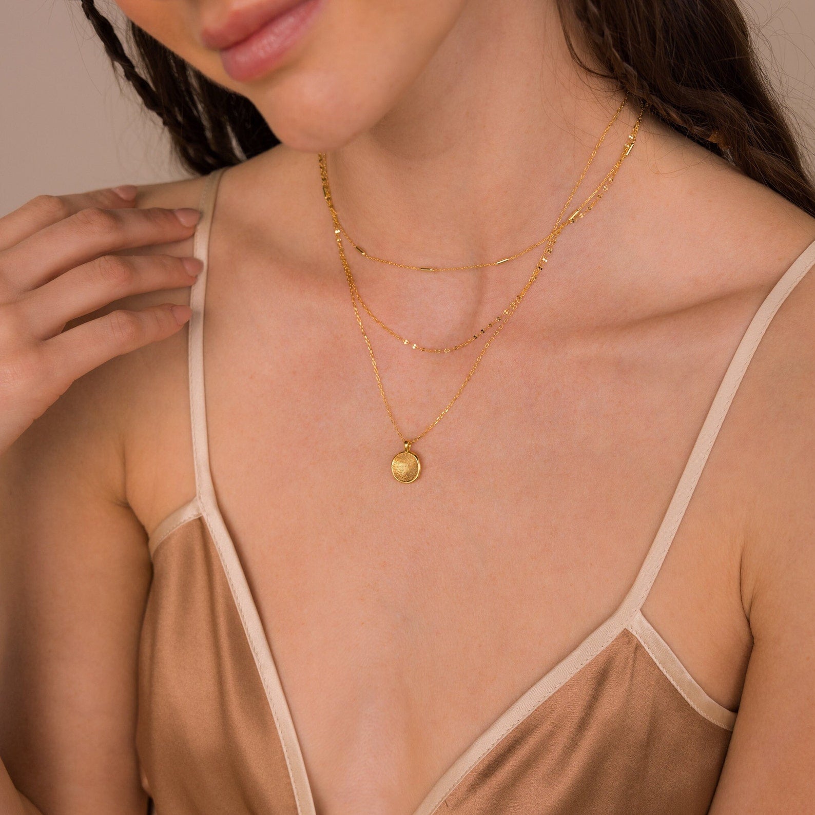 A woman wears a beige satin spaghetti strap top and layered gold necklaces, including the Fingerprint Coin Necklace, with her hand near her collarbone.