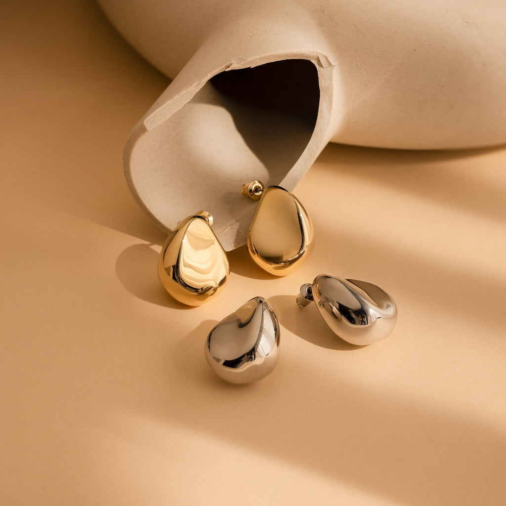 Bold Teardrop Studs in gold and silver rest on a beige surface with soft shadows, showcasing their geometric dome shape—perfect statement earrings for any look.