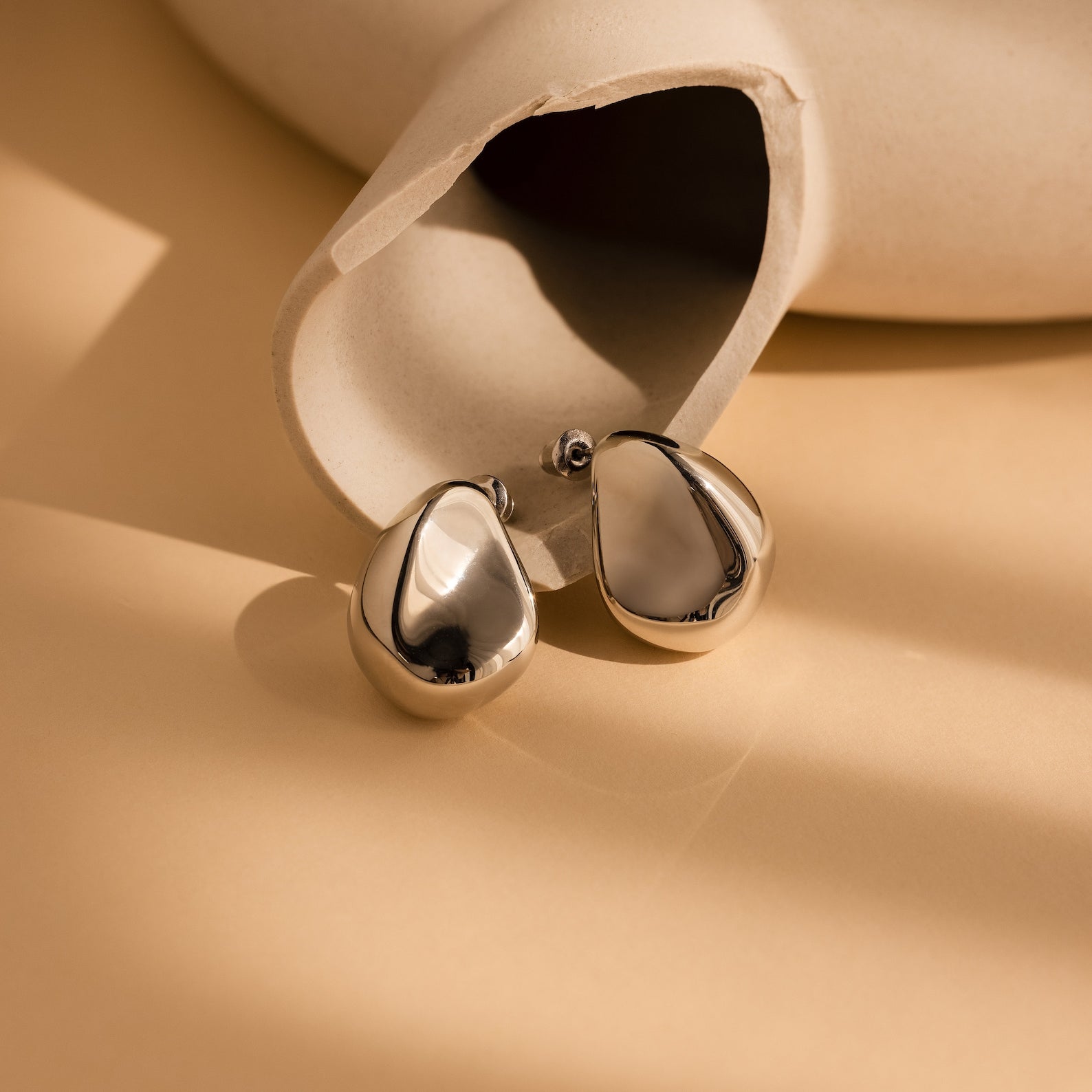 The Bold Teardrop Studs, featuring a shiny, eye-catching teardrop design, are displayed against a beige ceramic vase on a tan surface.