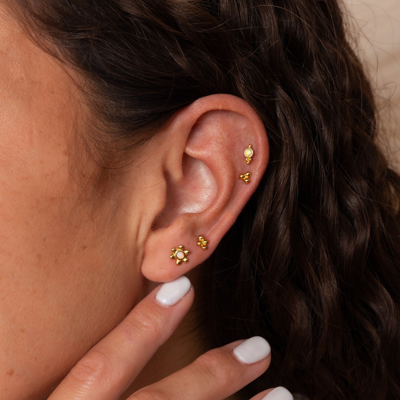 A close-up of an ear adorned with the Opal Cluster Studs Set and other gold earrings, as a hand with white nail polish gently touches the ear.