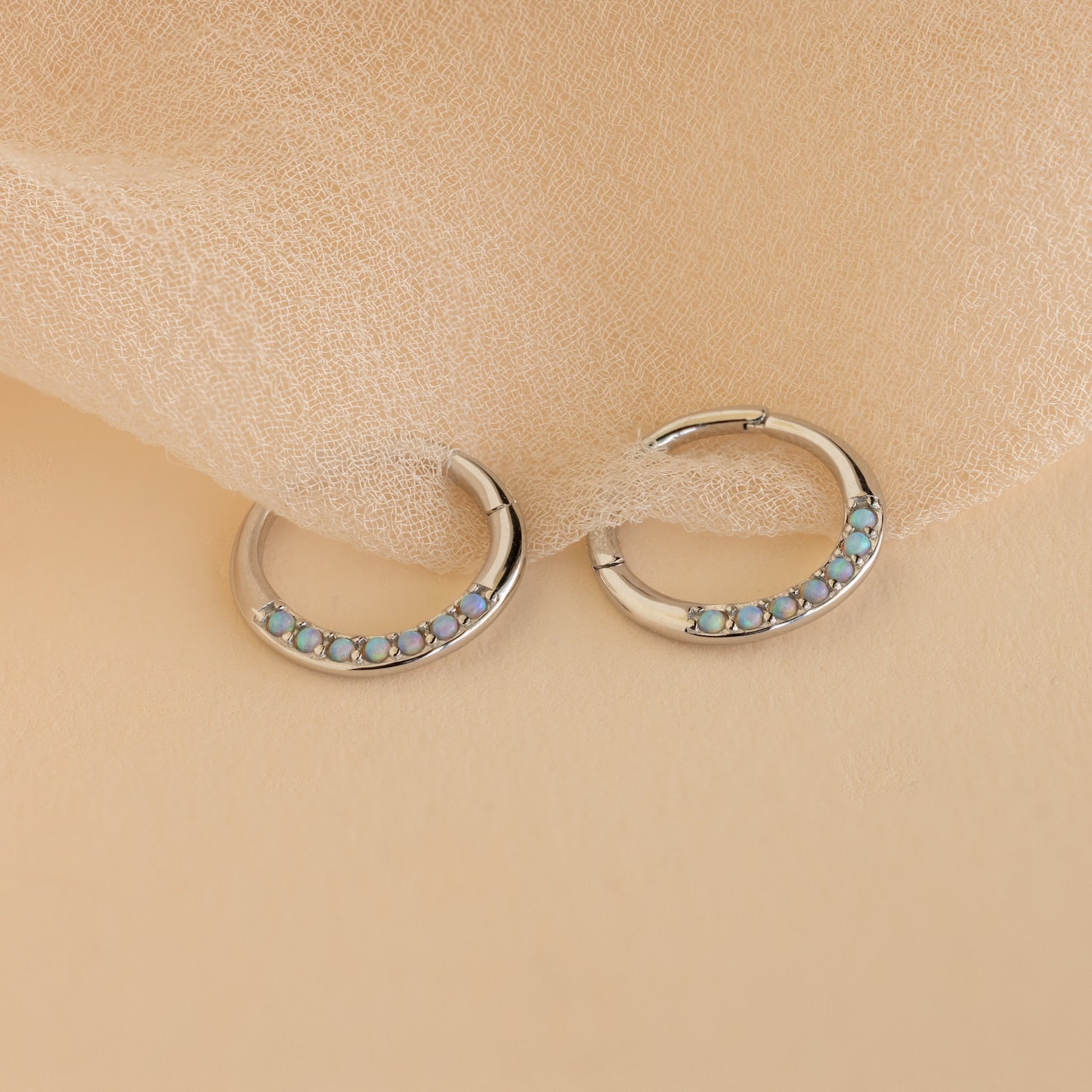 Opal Cartilage Huggies—silver hoop earrings with petite blue opals, ideal for minimalist style or as a chic helix piercing, displayed on a soft beige fabric background.