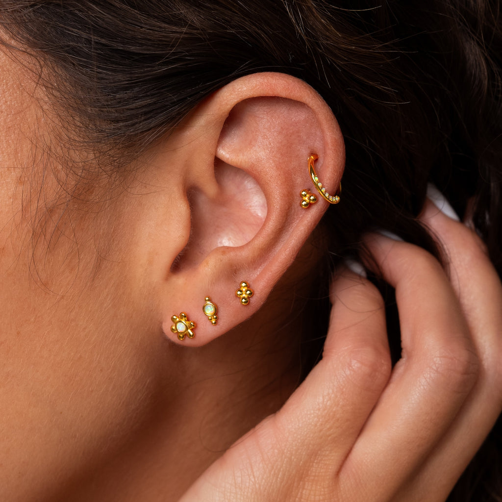 A close-up of an ear adorned with multiple gold earrings, featuring Opal Cartilage Huggies and minimalist styles, as a hand gently touches the side of the face.