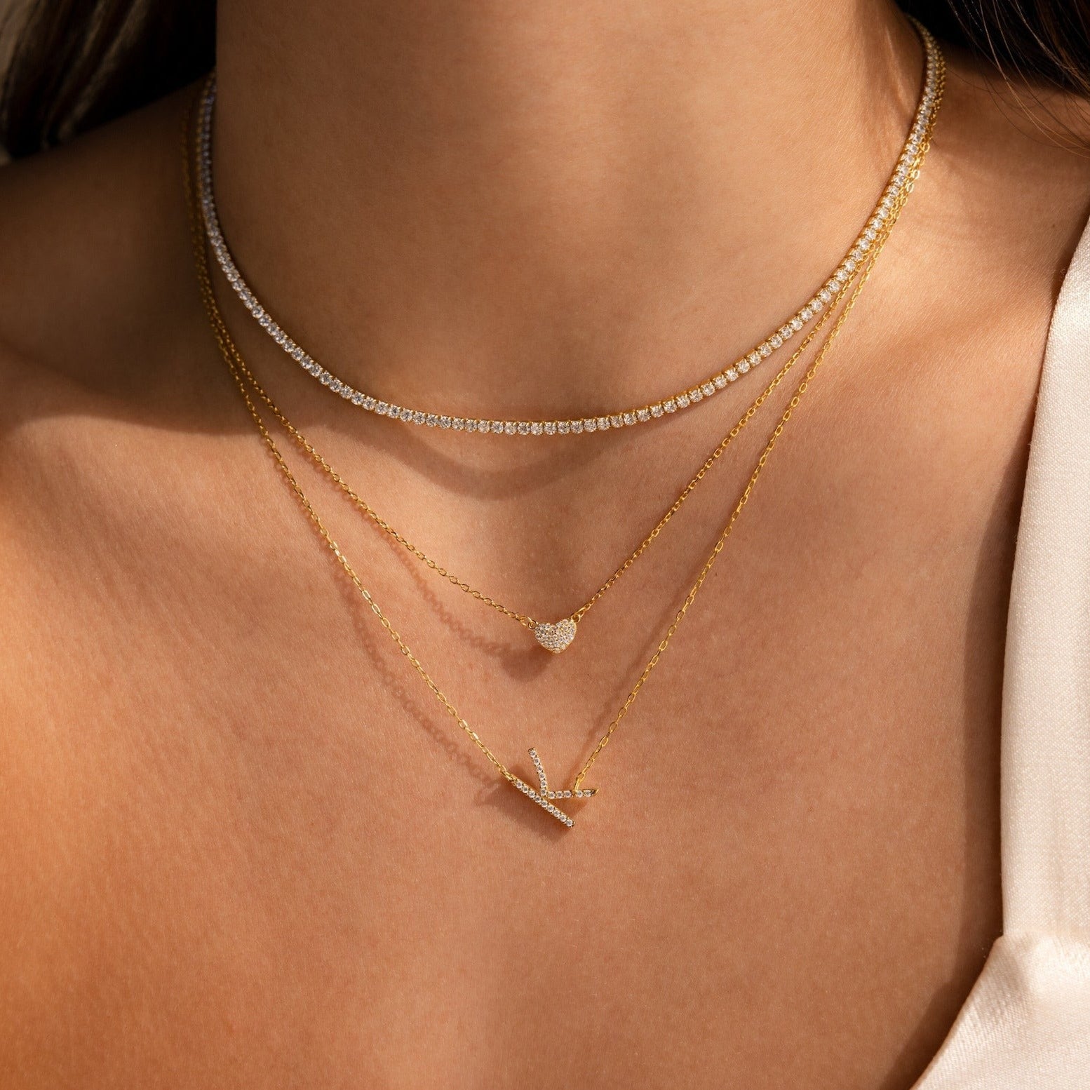 A woman wears a Mini Pave Sideways Initial Necklace—personalized, sparkling with gems—layered with a diamond bar and heart necklace for a beautiful, gold-accented look.