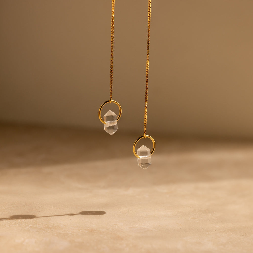 The Herkimer Diamond Threader Earrings, featuring two gold chains with clear crystal pendants, dangle gracefully against a neutral beige background.