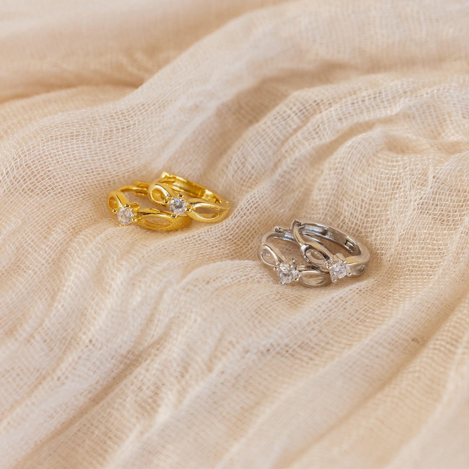 Diamond Infinity Hoops—elegant gold and silver earrings with sparkling diamonds in a romantic infinity design—are displayed on soft beige fabric.