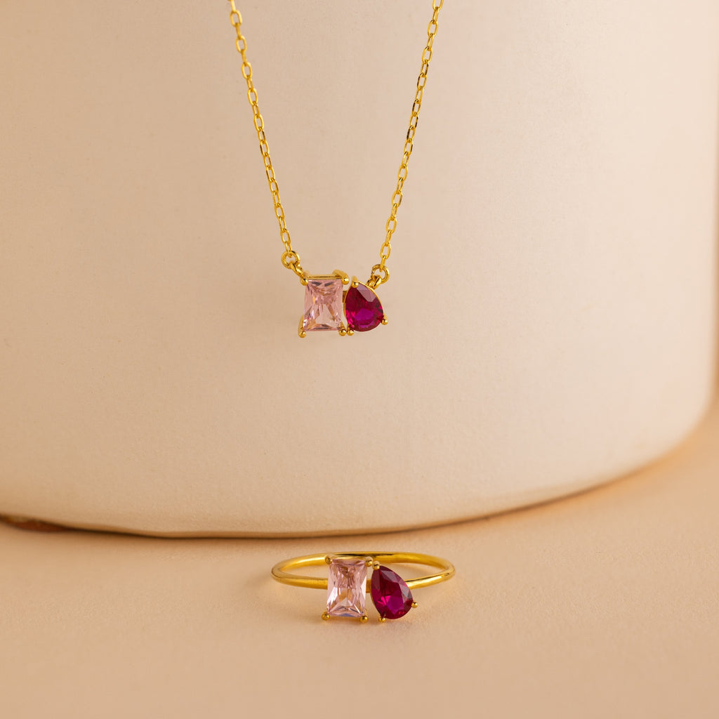 A gold necklace and the Toi et Moi Birthstone Ring in Rose Gold, featuring pink square and red teardrop gems, elegantly displayed on beige—a customizable ring that makes a thoughtful Christmas gift.