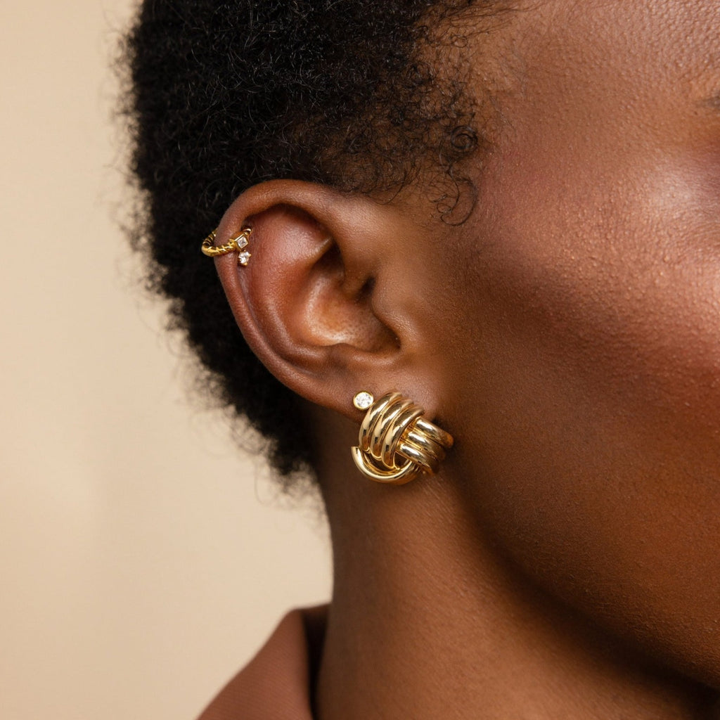 Close-up of a person’s ear adorned with multiple gold earrings, featuring hoops with a ribbed texture and the Bold Triple Knot Studs.