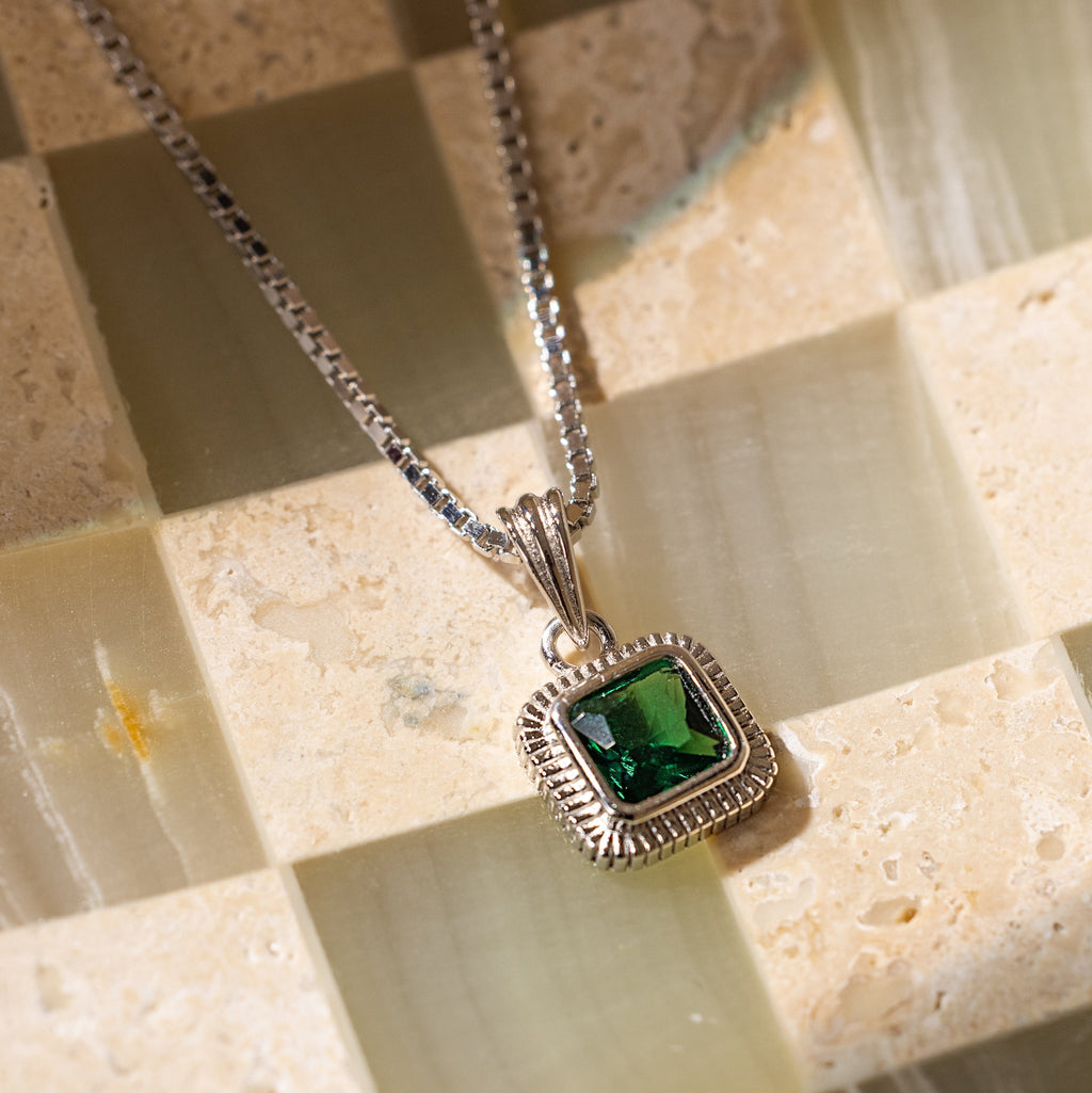 The Vintage Emerald Pendant Necklace showcases a square green gemstone on a silver chain, elegantly displayed on a checkered stone surface.