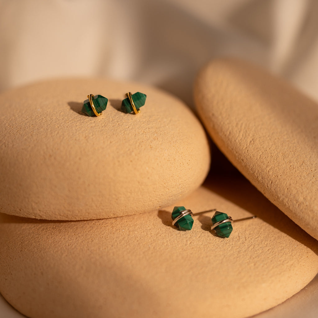 The Herkimer Malachite Studs feature green malachite with gold accents in a geometric design, elegantly displayed on smooth beige stones.