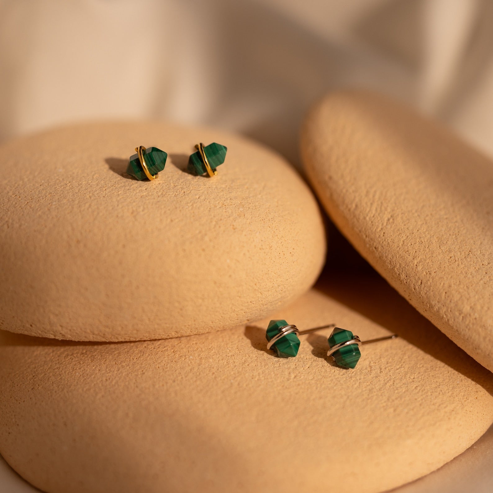 The Herkimer Malachite Studs feature green malachite with gold accents in a geometric design, elegantly displayed on smooth beige stones.