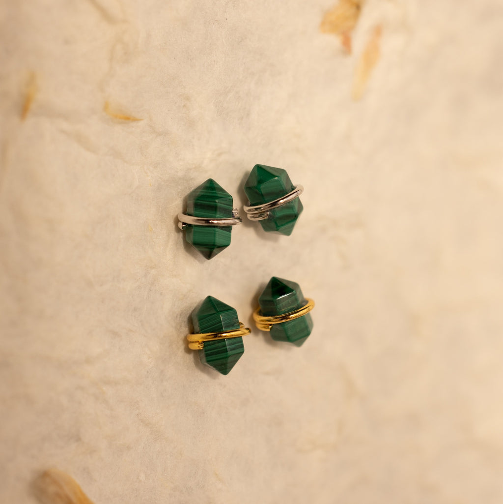 The Herkimer Malachite Studs feature geometric green malachite stones with gold and silver bands, displayed on a textured beige surface.
