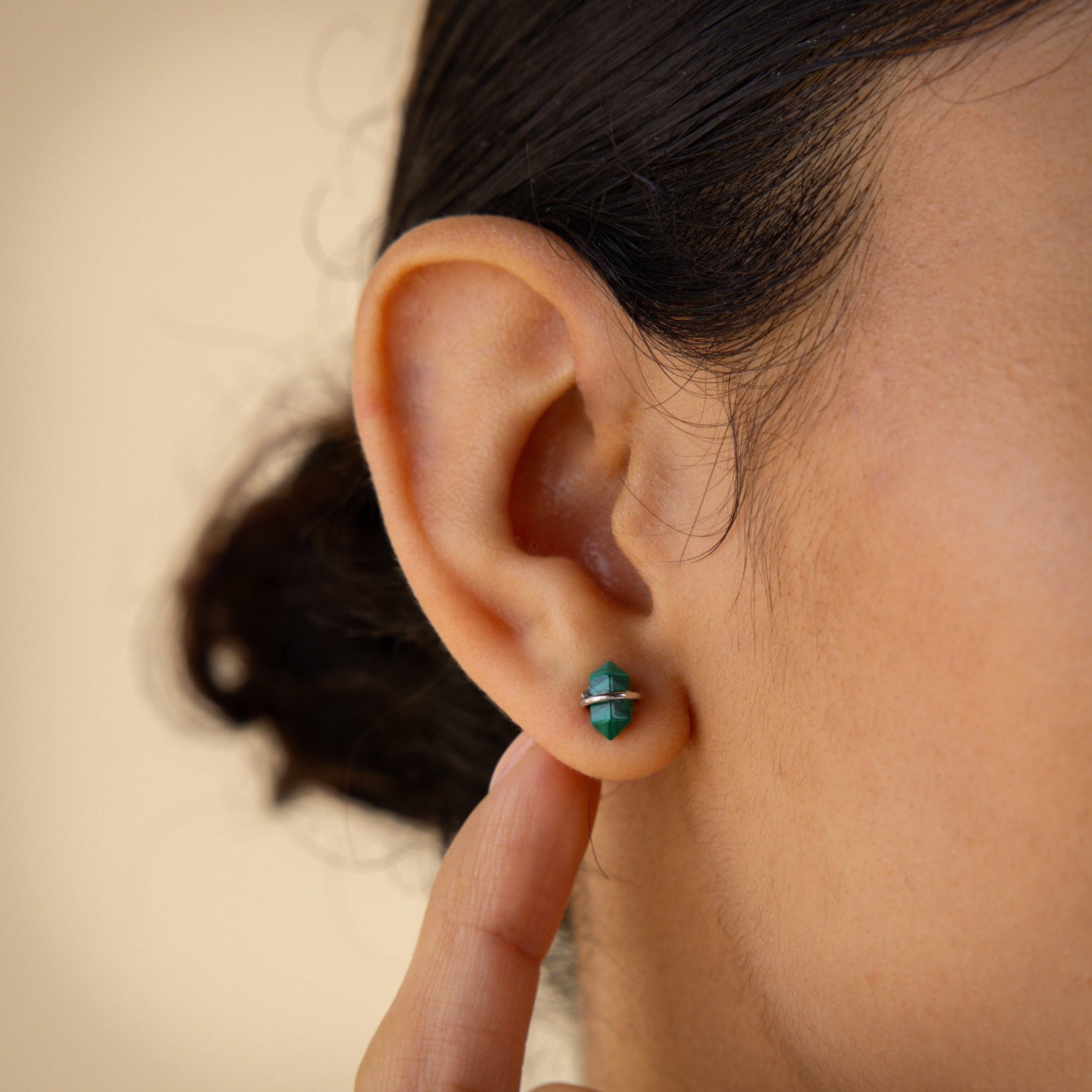 Someone touches their ear while wearing the Herkimer Malachite Studs, small earrings featuring green and silver accents.
