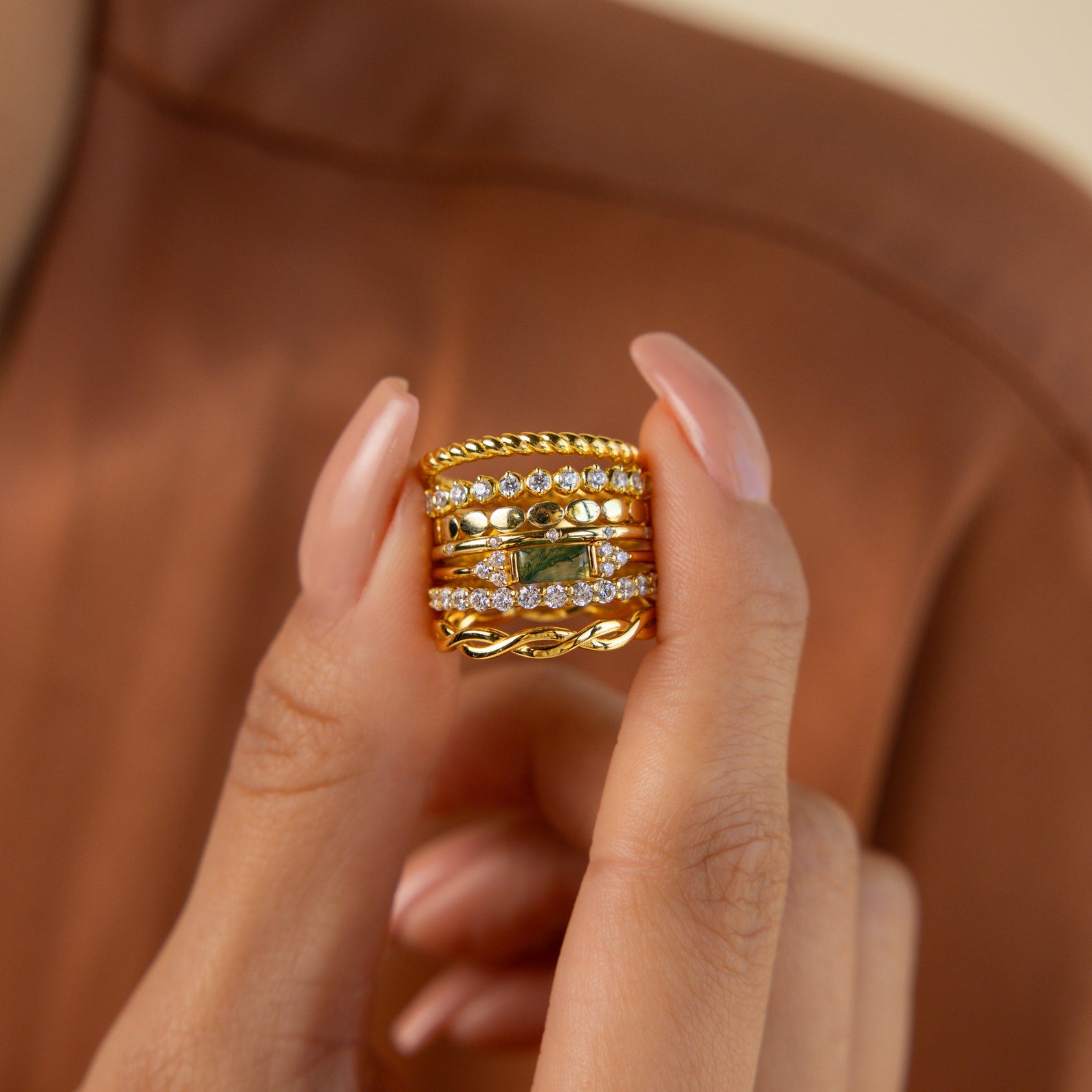 A manicured hand holds a stack of gold and diamond rings, including the Agate Baguette Ring, set against a brown background.