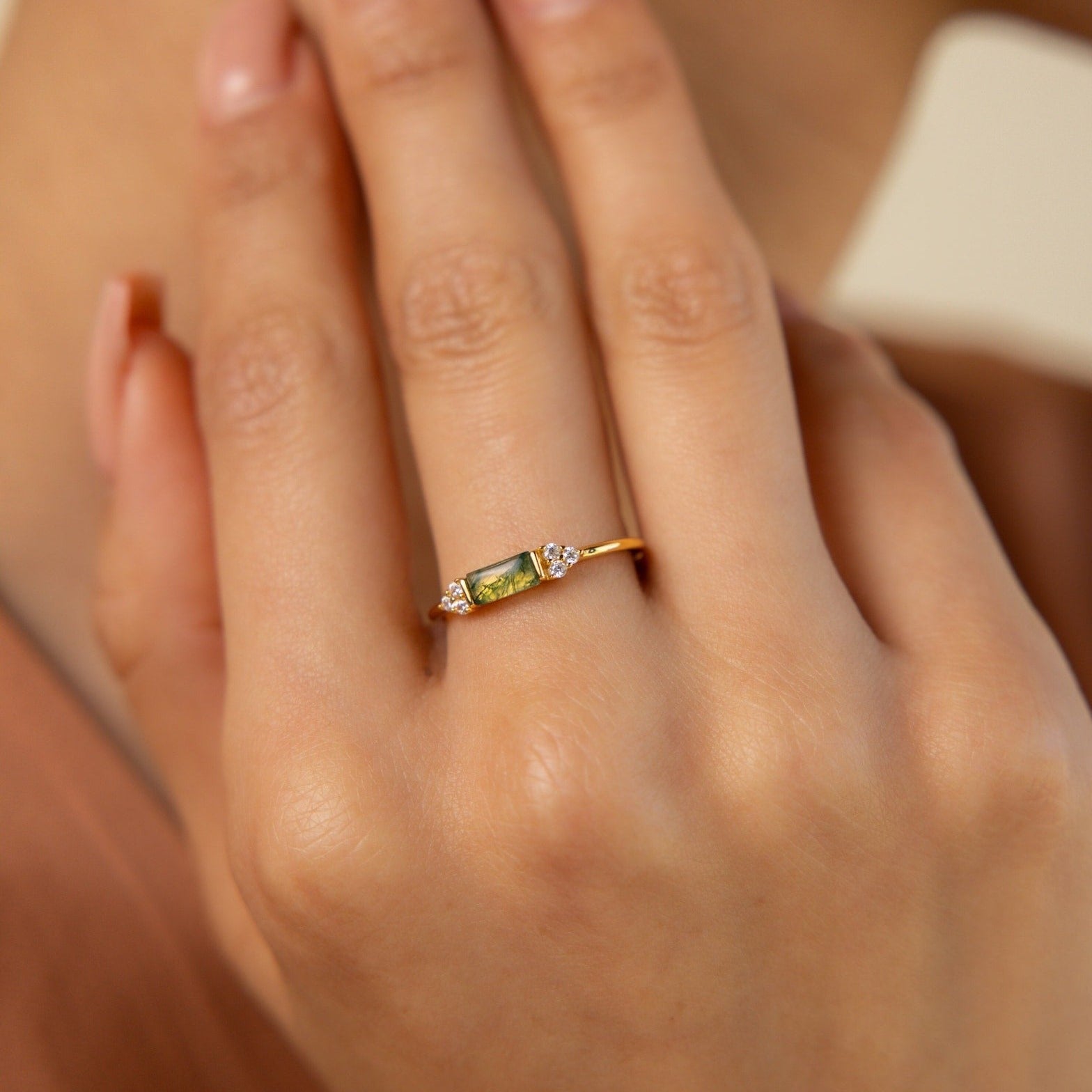 A hand wears the Agate Baguette Ring, featuring a gold band with a green gemstone and two small clear stones, as fingers gently touch the face.