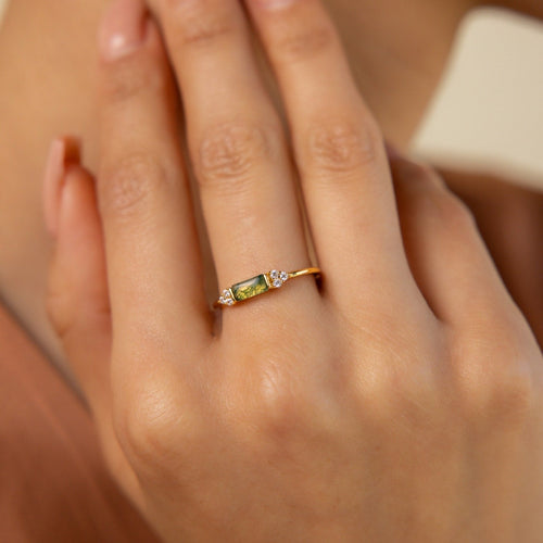 A hand wears the Agate Baguette Ring, featuring a gold band with a green gemstone and two small clear stones, as fingers gently touch the face.