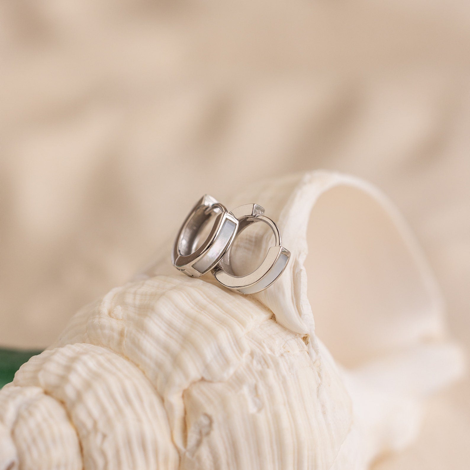 The Mother of Pearl Inlay Huggies are displayed on a textured white seashell set against a soft beige background.