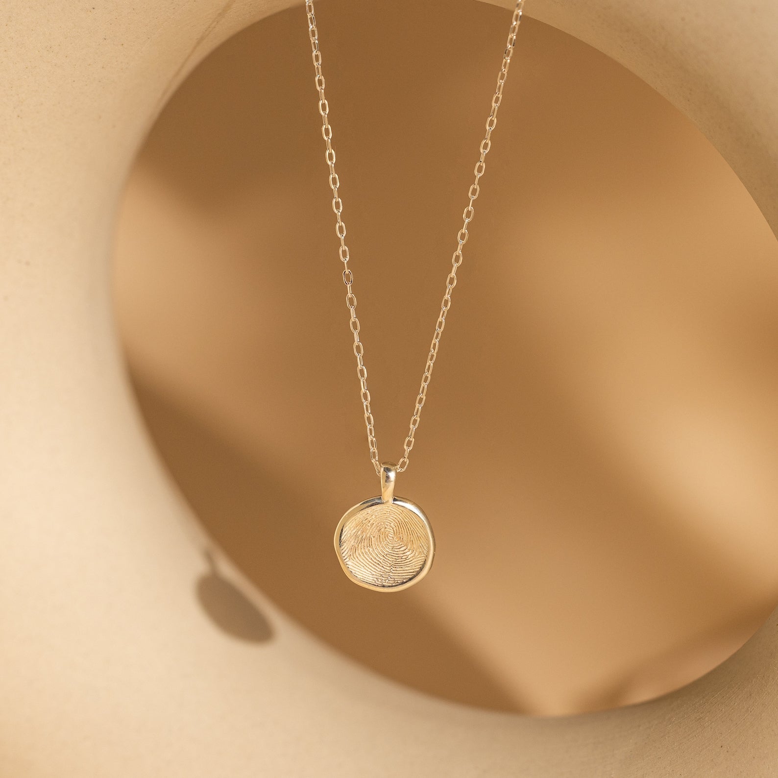 The Fingerprint Coin Necklace features a round gold pendant engraved with a unique fingerprint, shown on a soft beige background—an elegant and meaningful gift, perfect for Mother's Day.