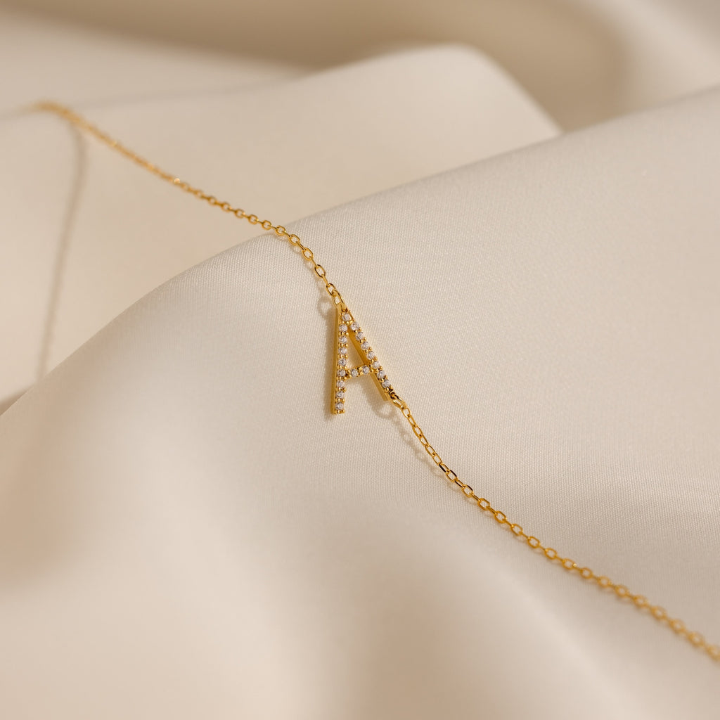 The Mini Pave Sideways Initial Necklace features a gold letter A pendant displayed on soft, cream-colored fabric.