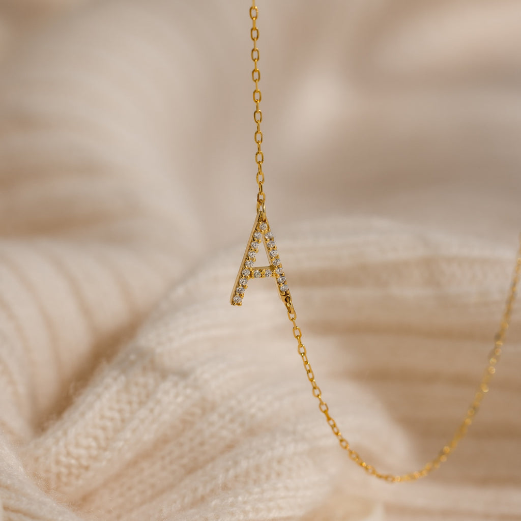 The Mini Pave Sideways Initial Necklace features a diamond-studded letter A pendant and is displayed on a cream knit fabric background.