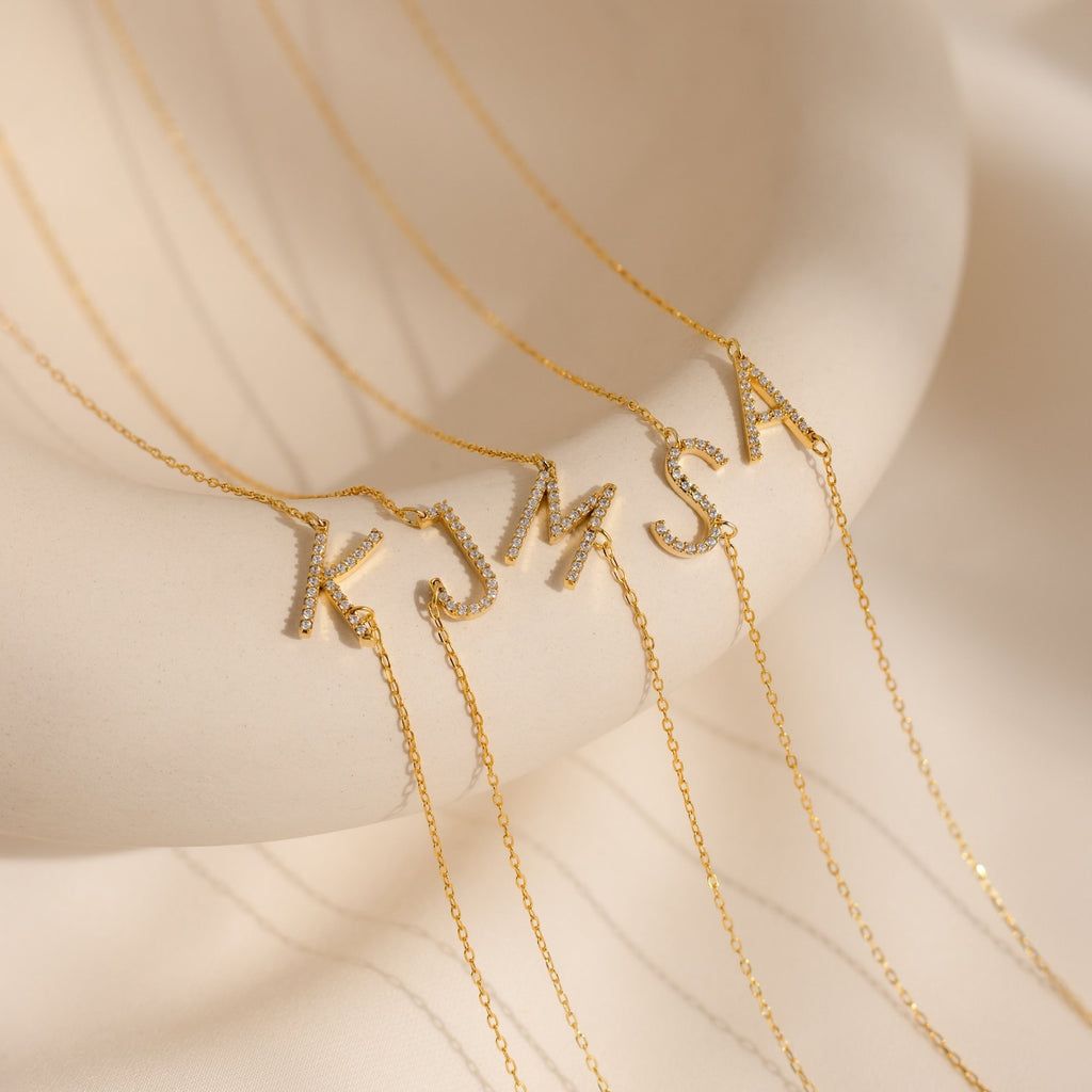 Sparkling gold Mini Pave Sideways Initial Necklaces in letters K, J, M, and S are showcased on a smooth cream display—perfect for personalizing your look.