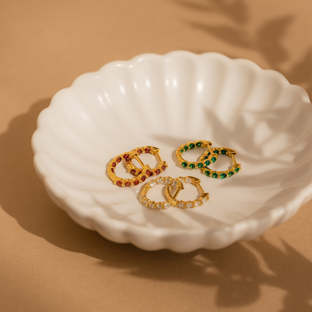 The Inside Out Emerald Huggies, along with three pairs of gold hoop earrings featuring red, green, and clear stones, are displayed on a white scalloped dish—ideal for fans of colorful birthstone jewelry.