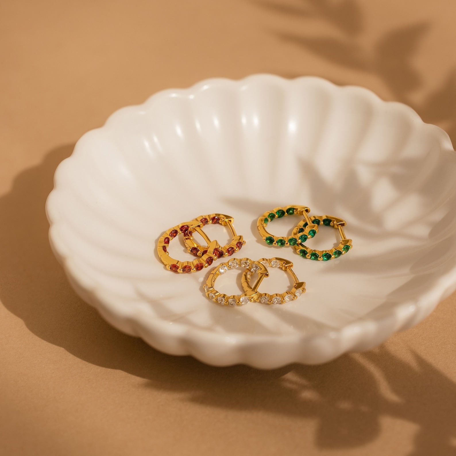 The Inside Out Emerald Huggies, along with three pairs of gold hoop earrings featuring red, green, and clear stones, are displayed on a white scalloped dish—ideal for fans of colorful birthstone jewelry.